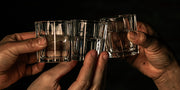 Three hands holding mezcal in small glasses on a dark background